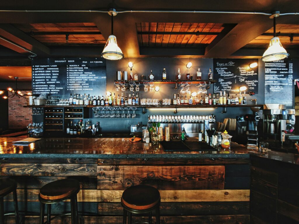 Wnętrze stylowego baru z drewnianym blatem, dwoma stołkami barowymi i półkami z butelkami alkoholu, oświetlone ciepłym światłem lamp, z tablicami menu na ścianach.